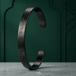 Black stainless steel cuff bracelet standing upright on a dark green background.